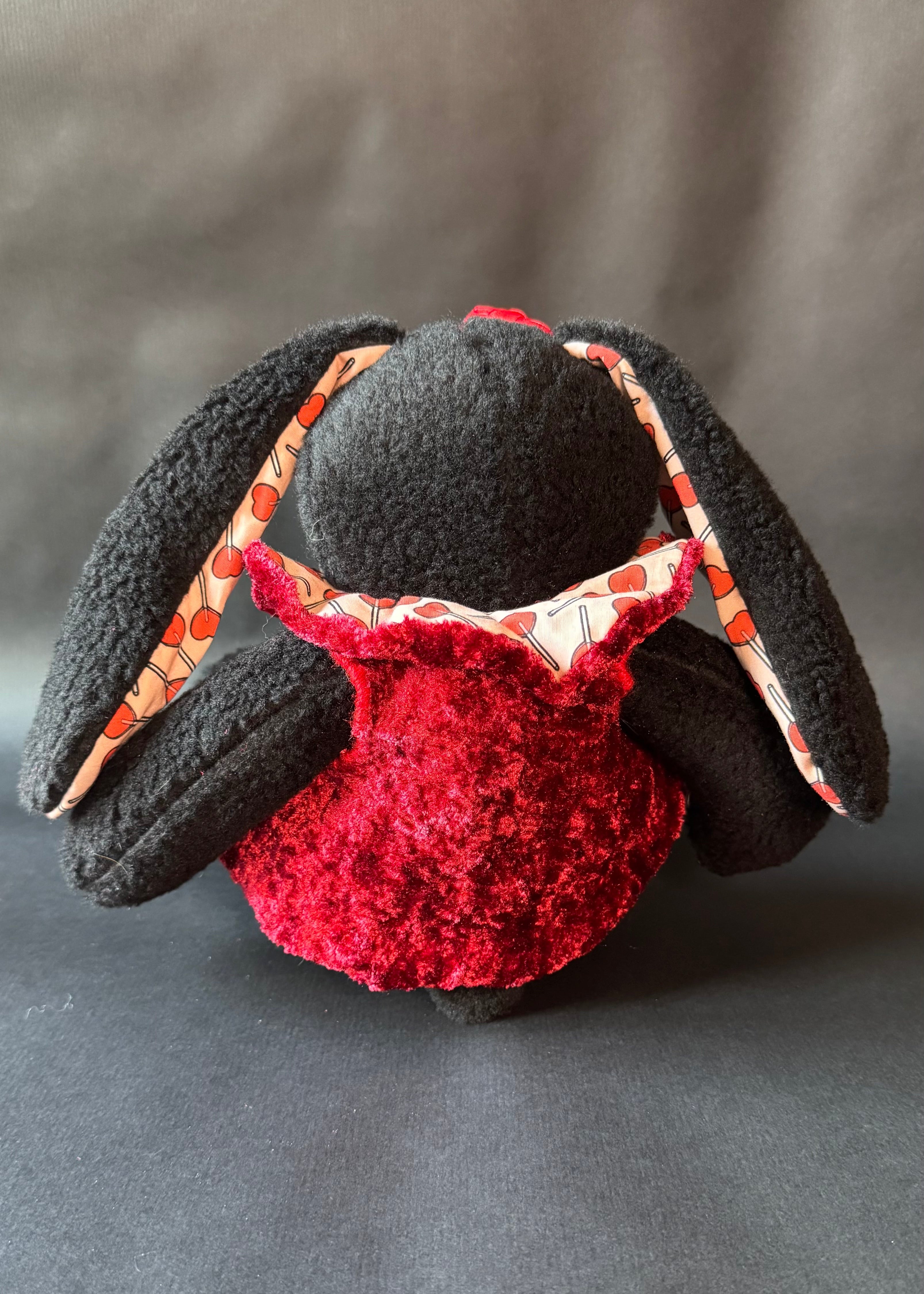 Das Bild der Rückseite eines schwarzen Kuscheltier Hasen mit Herzlolli-Stoff-Highlights und roter Jacke an.